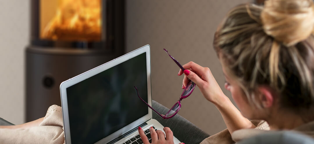 Dame med hårtopp sitter foran en sort vedovn Scan 67 med PC i fanget og briller i hånden