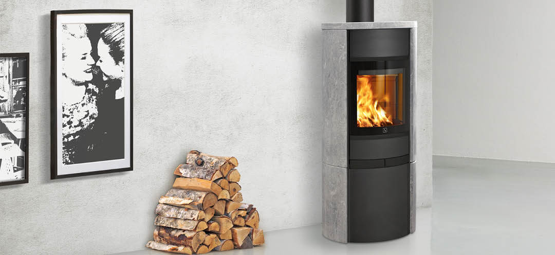 Sort vedovn med klebersteinssider Scan 68-15 i hvitt rom med en vedstabel plassert ved siden