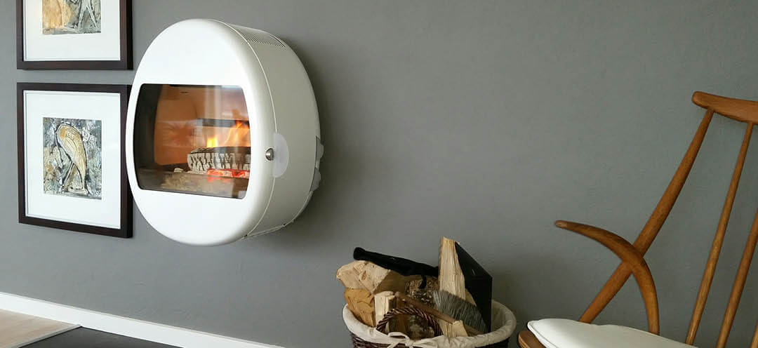 Hvit vegghengt vedovn Scan 66-1 med eggeform, henger på veggen i en grå stue med en vedkurv og stol ved siden av