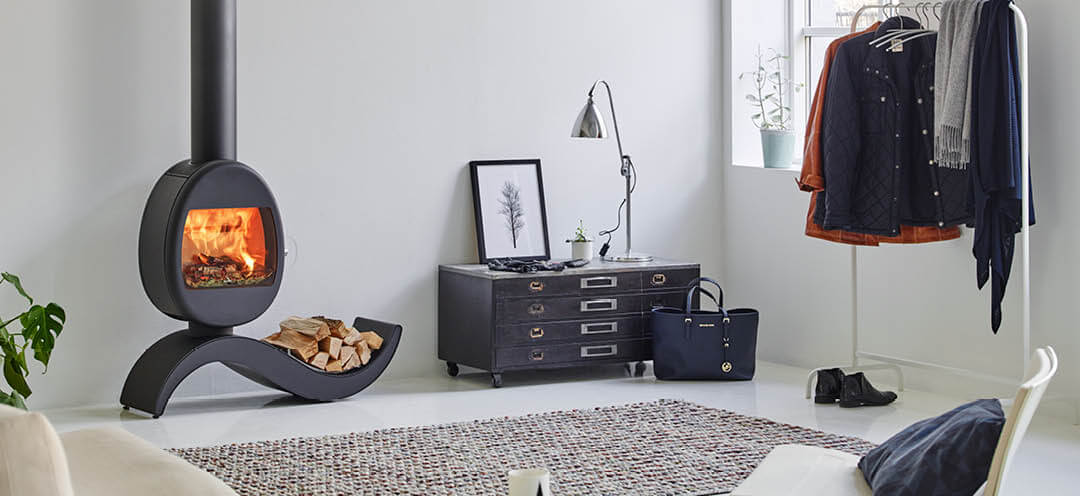 Sort vedovn Scan 66-5 med s-formet base med vedholder, i en liten hvit leilighet med sort kommode og klesstativ med klær