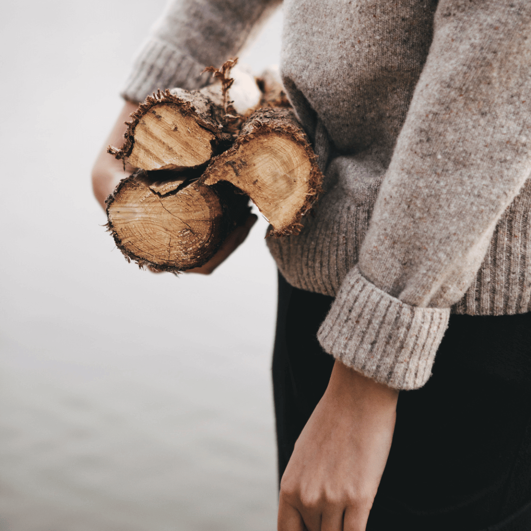 Person kledd i ullgenser bærer vedkubber i armene, med rolig vann i bakgrunnen