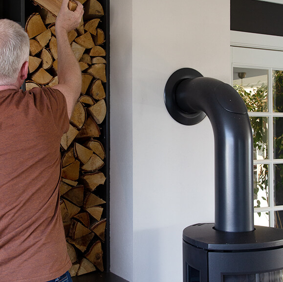 Sort vedovn med knerør inn i hvit pipe. Mann i brun t-skjorte legger inn ved i vednisje.