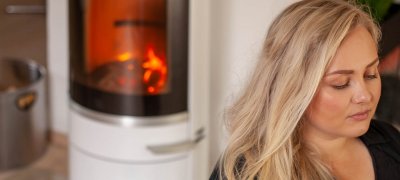 Blond kvinne med langt hår sitter ved en hvit vedovn Scan 83-2 som brenner