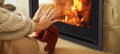 Person med varme klær og ullsokker varmer hendene foran en peisovn med synlige flammer – illustrasjon av trygg og koselig oppvarming i krisesituasjoner