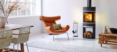 Sort oval vedovn med aluminiumsdetaljer og vedholder i basen står i en lyst stue med en orange skinnstol og spisebord ved siden av