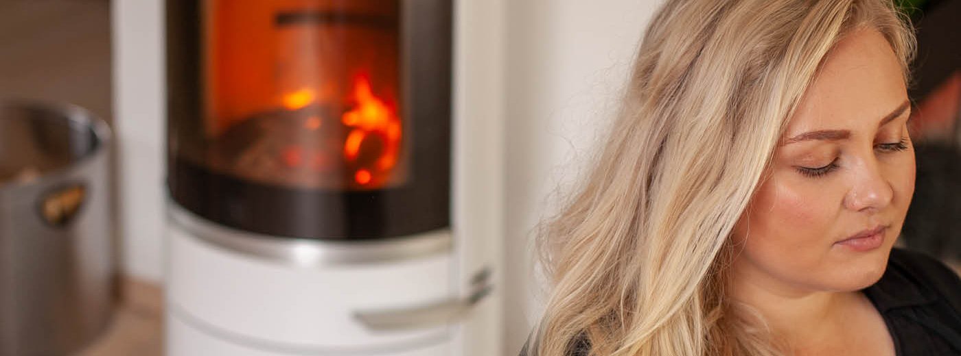 Blond kvinne med langt hår sitter ved en hvit vedovn Scan 83-2 som brenner