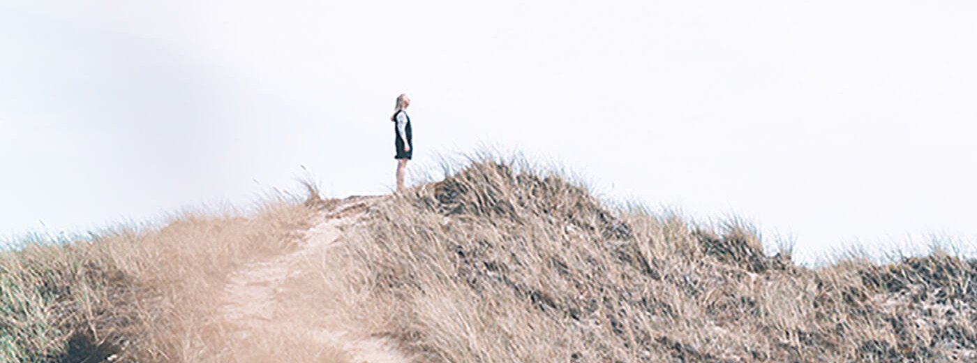 Jente med blå kjole står på en dansk sandklippe og nyter vinden i håret