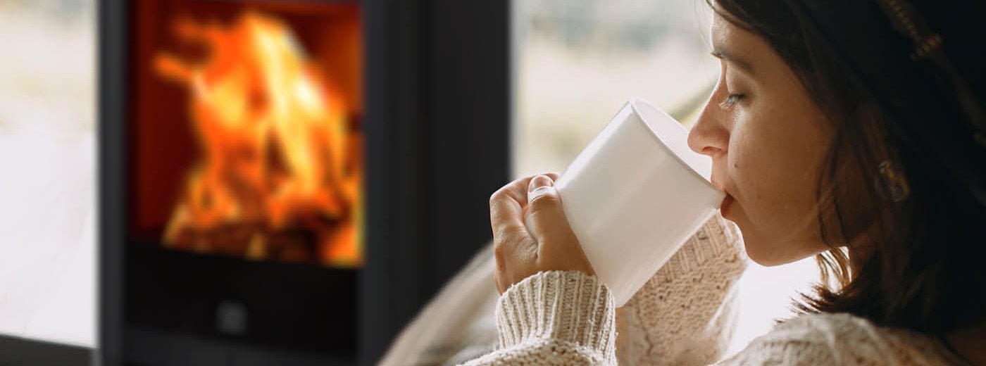 Jente med mørkt hår og hvit ullgenser drikker av hvit kopp foran vedovn Scan 65-1