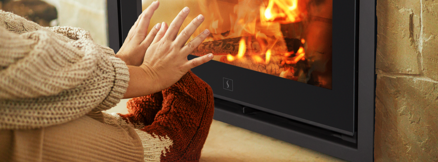 Person med varme klær og ullsokker varmer hendene foran en peisovn med synlige flammer – illustrasjon av trygg og koselig oppvarming i krisesituasjoner
