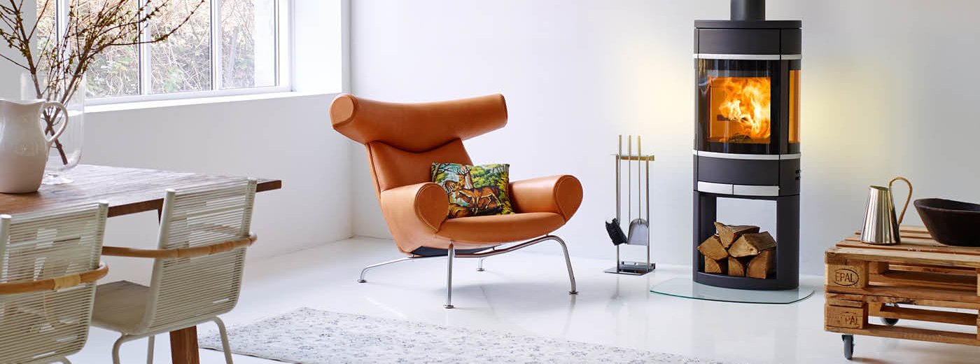 Sort oval vedovn med aluminiumsdetaljer og vedholder i basen står i en lyst stue med en orange skinnstol og spisebord ved siden av