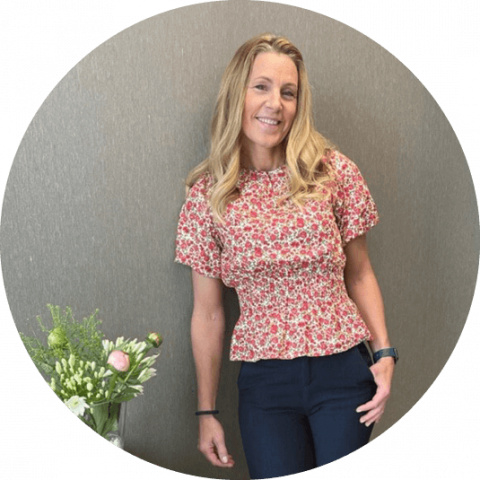 Blond kvinne i blomstrete topp står lent inntil en beige vegg